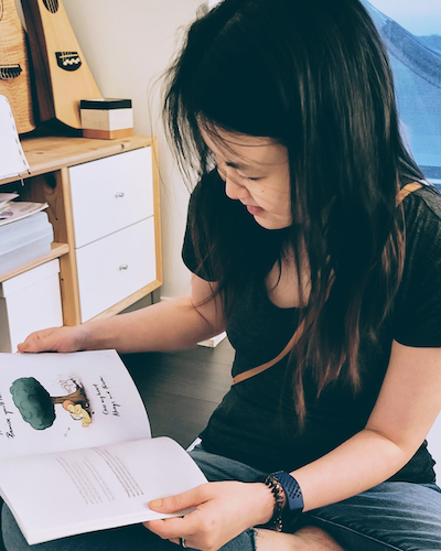 "A close-up view of Viola Hernandez sitting on the floor and looking down as she reads an open illustrated children's book. In the background, wooden shelves hold small string instruments and storage boxes."
