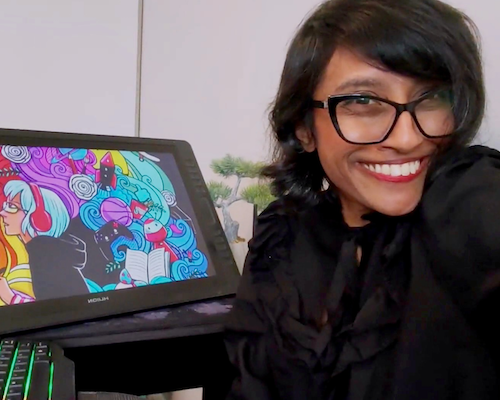 "A selfie of Su Irugalratne smiling while wearing black-rimmed glasses, sitting beside a large drawing tablet that displays her colorful digital artwork."