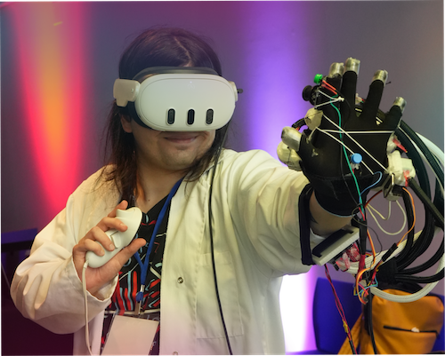 "A photograph of Milsey wearing a white VR headset and a white lab coat. They are holding a white controller in one hand and extending their other hand forward, which is wearing a black glove rigged with complex wires and sensors. The background features soft purple and pink stage lighting."