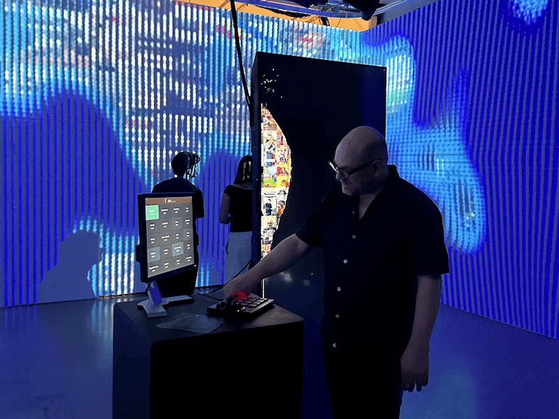 "A man in glasses and a black short-sleeved shirt operating a digital control pad on a podium beside a computer monitor. In the background, large blue LED screens illuminate an immersive installation space as other visitors look on."