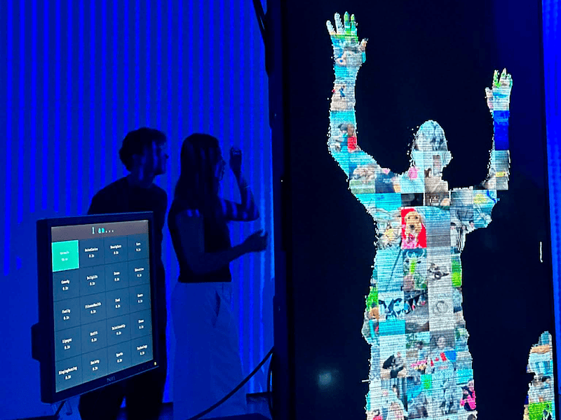 "A large vertical digital screen displaying a person's pixelated silhouette made of various small photos, with hands raised in the air. To the left, two people stand in silhouette beside an interactive monitor in a room lit by vibrant blue LED lights."