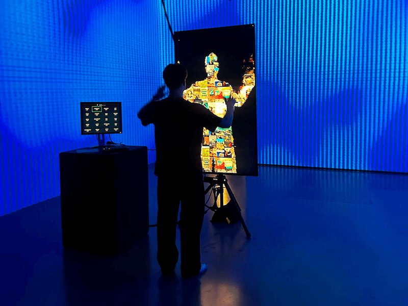 "A person in silhouette facing a vertical display screen, raising their hands to interact with a photo-mosaic figure on the monitor. The room is surrounded by large, bright blue LED walls, with an interactive control station positioned to the left."