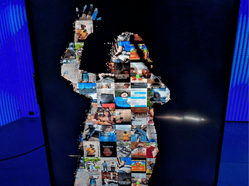 "A close-up view of a vertical digital display showing a human silhouette composed of a mosaic of small, varied photographs. The figure appears with one hand raised, set against a dark background flanked by blue LED panel lights."