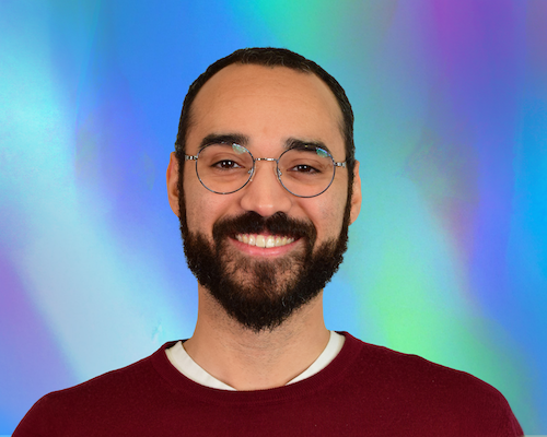 "A portrait of Ryan Boyd smiling with round wire-rimmed glasses and a dark beard, wearing a maroon crewneck sweater over a white shirt. The background features a vibrant blue, purple, and green gradient overlay."