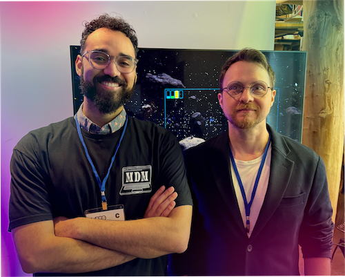 "A portrait of Ryan Boyd and Konrad Karczewski standing side-by-side indoors. Ryan Boyd, on the left, is smiling with his arms crossed and wearing an 'MDM' t-shirt, glasses, and an event lanyard. Konrad Karczewski, on the right, wears round glasses, a dark blazer, and an identical lanyard. A monitor displaying a space or asteroid video game screen is behind them, accented by subtle pink and yellow gradient lighting on the outer edges."