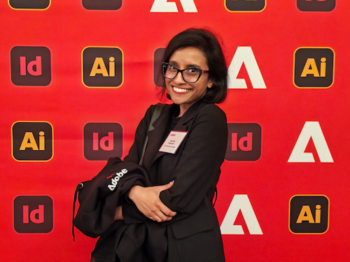 A portrait of a smiling woman with glasses and dark shoulder-length hair wearing a black blazer with a name tag, holding a black jacket branded 'Adobe'. She poses in front of a red step-and-repeat backdrop featuring Adobe logos and software icons such as Illustrator and InDesign."