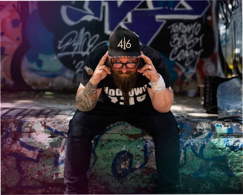 "A photograph of Alex Sein sitting on a concrete ledge in a location covered in colorful graffiti. He is leaning forward with his hands pointing to his temples, looking directly into the camera. He has a long, groomed beard, glasses, a tattooed arm, and is wearing a black '416' baseball cap and a black graphic t-shirt. The background features various street art tags, including the words 'Toronto Beast Coast.' The image has a soft pink and purple color cast on the left side."
