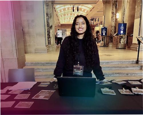 "Alisha Pereira, a Master of Digital Media student, smiles while standing behind a registration table at the Royal Ontario Museum (ROM)."