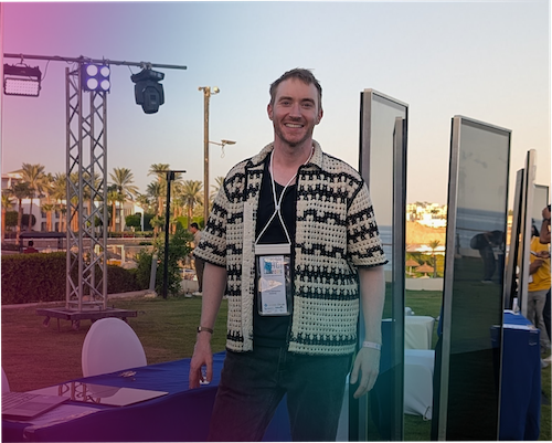 "Person standing in front of presentation table at an outdoor conference with a poster to the left. Pink, purple, and blue gradiaent overlay." 