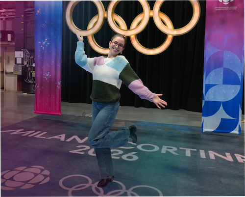 "A smiling woma n 'Iliana Mehmeti' wearing a colour-blocked sweater and glasses strikes a playful pose on a stage. Behind her is a large, illuminated Olympic rings logo flanked by CBC Olympic broadcasting banners. The stage floor features the CBC logo, the Olympic rings, and text that reads 'MILANO CORTINA 2026'."