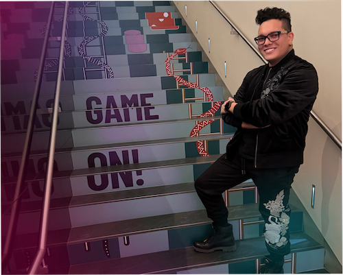 "A full-body photograph of Michael Carter-Arlt standing on an indoor staircase. He is smiling with his arms crossed, wearing black glasses, a black bomber jacket with floral embroidery on the shoulder, and black joggers featuring a large white floral design on the lower leg. The stairs are decorated with a 'Snakes and Ladders' game theme and the words 'GAME ON!' printed in purple. The left side of the image has a pink-to-purple transparent color overlay."