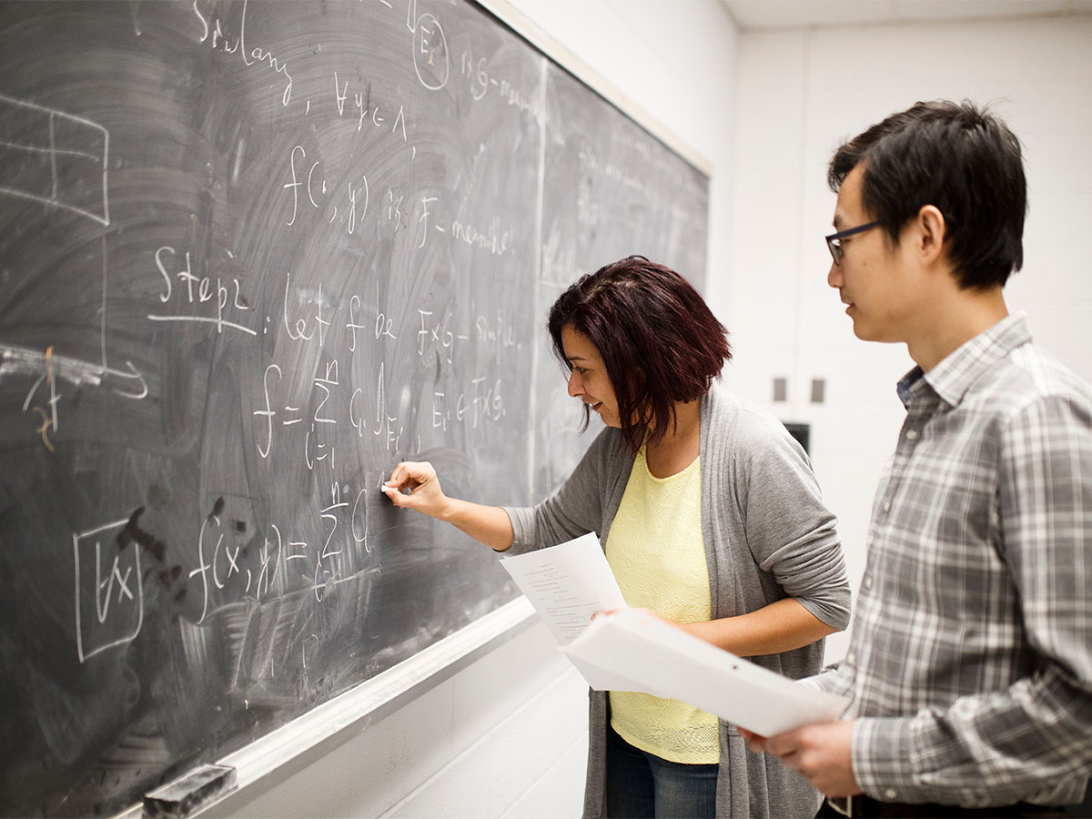 Student smiling while writing an equation on a black board while the instructor looks at it 