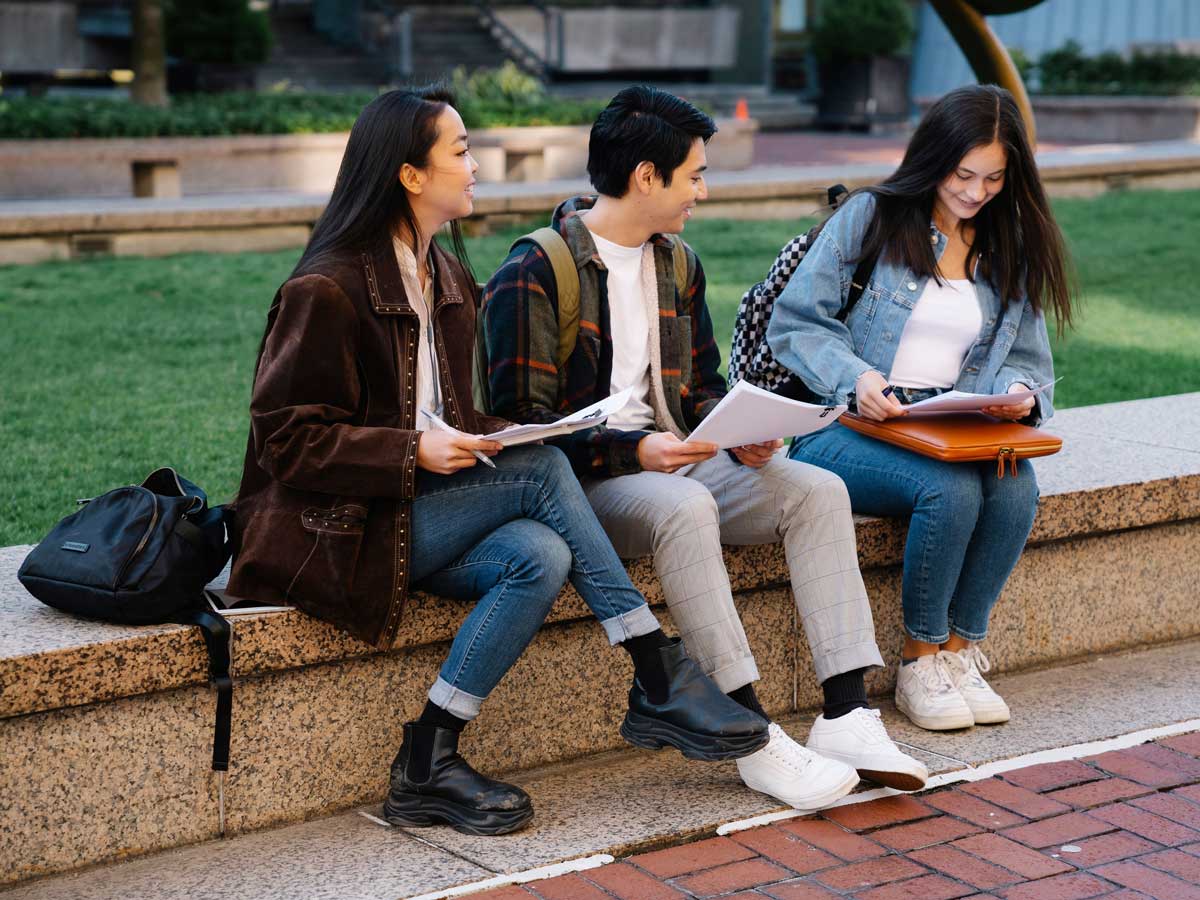 A group of three students sitting outdoors while reviewing papers together.