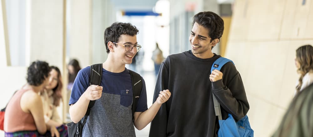 Two students walk through a university hallway while talking and smiling. Other students are visible in the background, creating a welcoming and active campus environment.