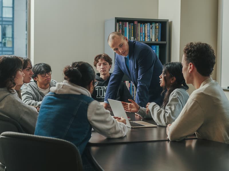 Students and a faculty member gathered around a meeting table in a bright classroom or study space. The group is engaged in discussion, with laptops open and participants actively listening and sharing ideas.