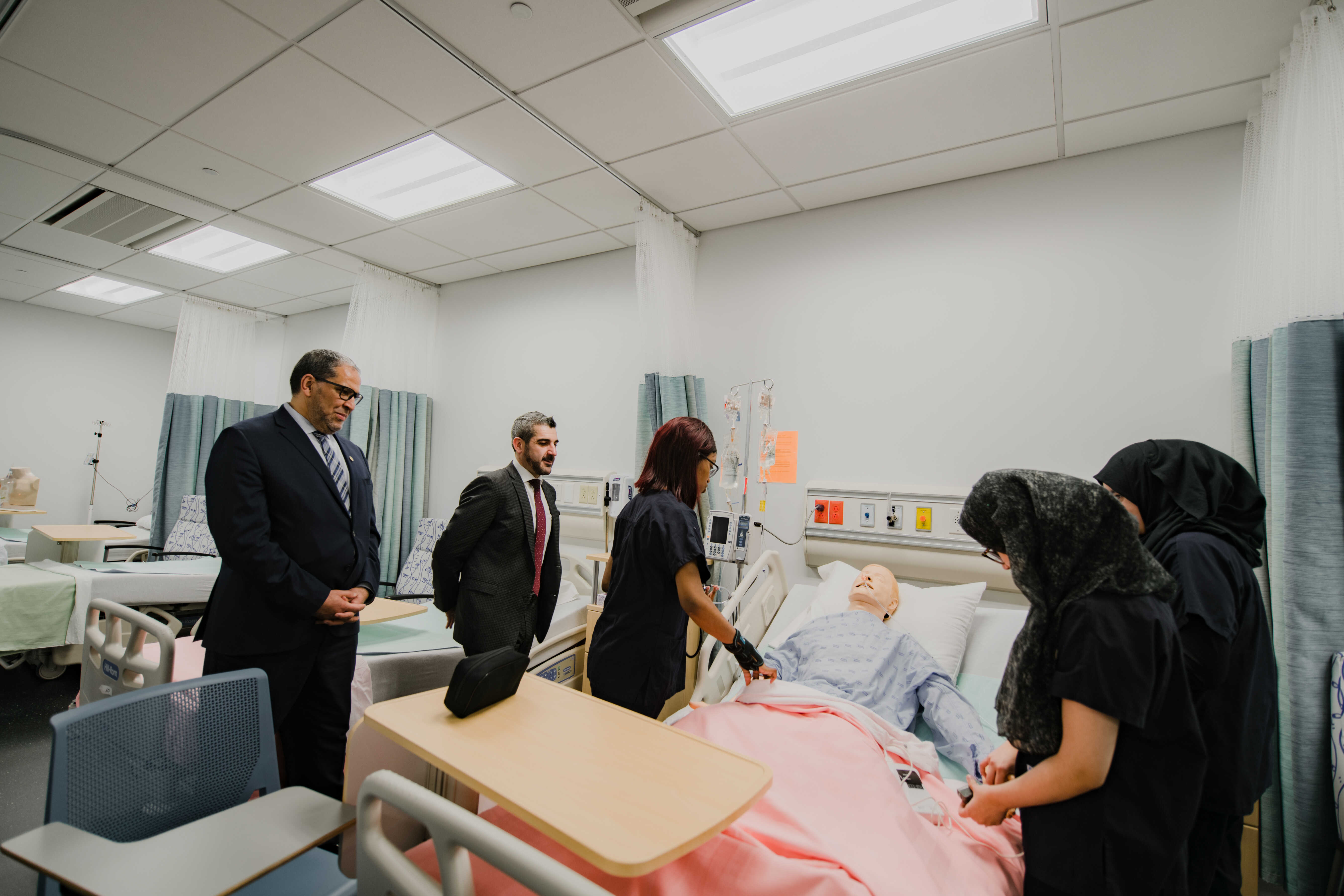 Students demonstrate clinical training techniques using a patient mannequin in a healthcare simulation room. Visitors observe the presentation and learn about hands-on nursing and healthcare education.