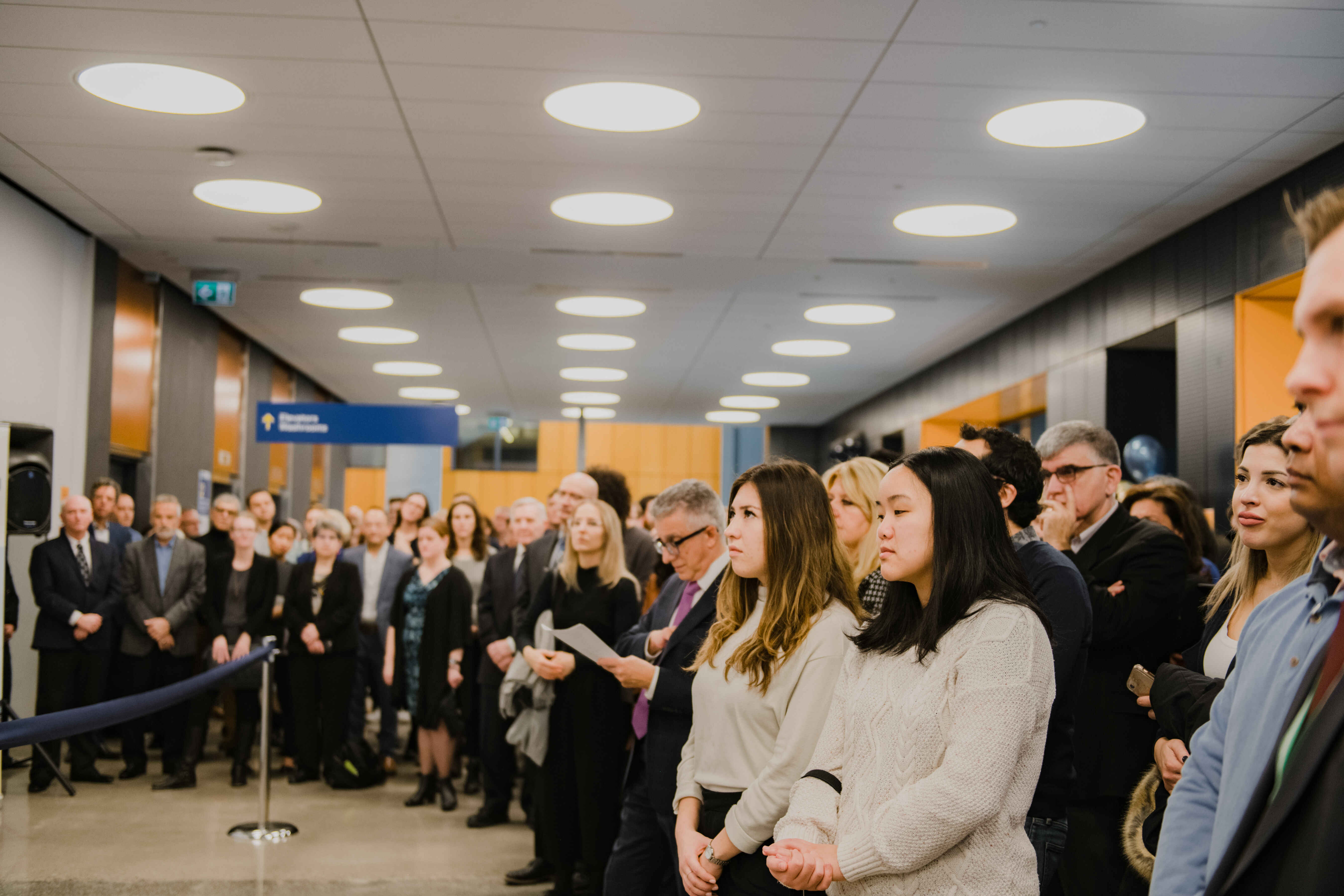 Students, faculty, staff, and guests stand together at a university event, listening attentively during a ceremony or announcement. Attendees fill the hallway and gathering space, creating a formal and engaged atmosphere.