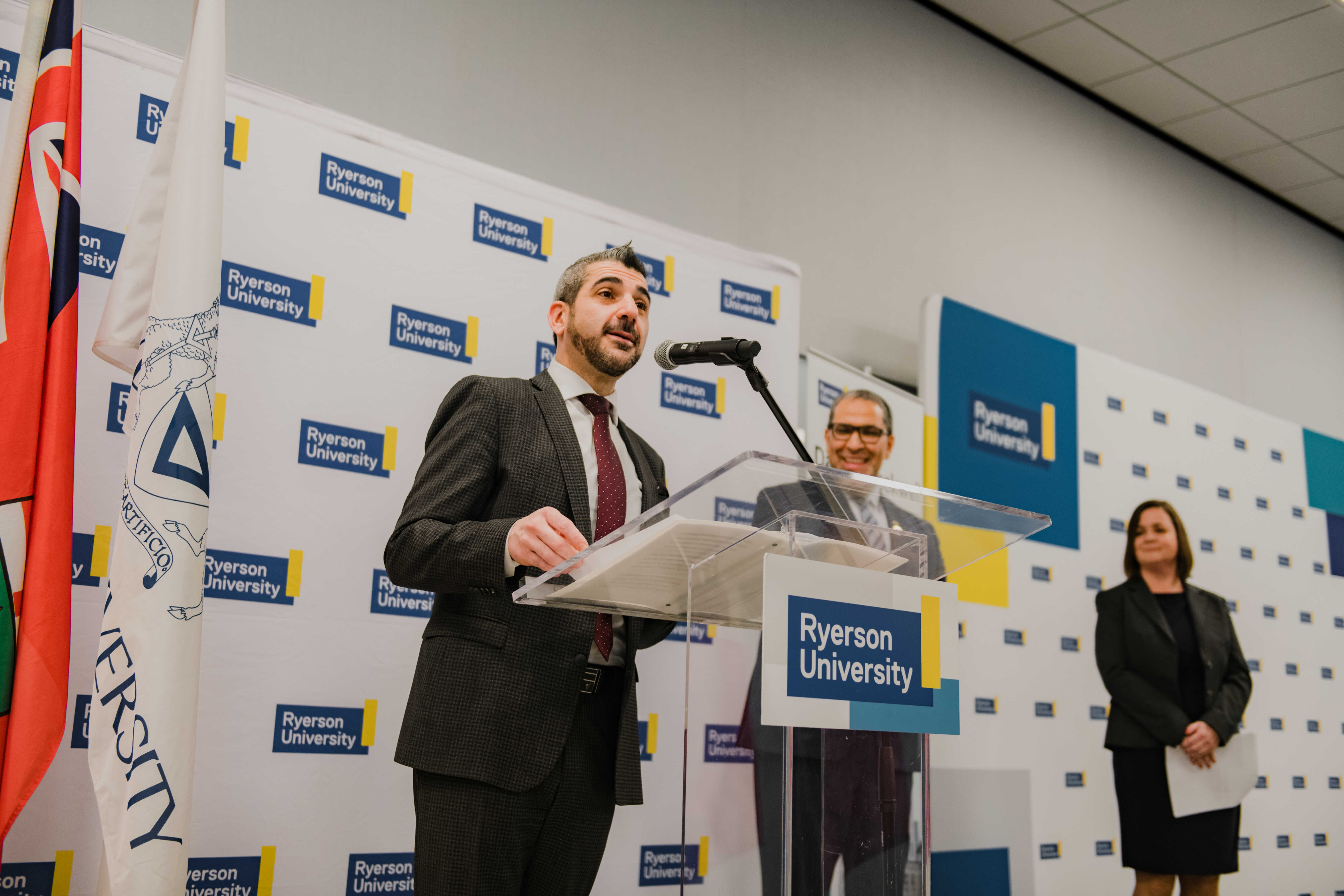 An individual speaks at a podium during an official university event while attendees and guests look on. Event signage and a crowd gathered nearby suggest a formal announcement or opening ceremony.