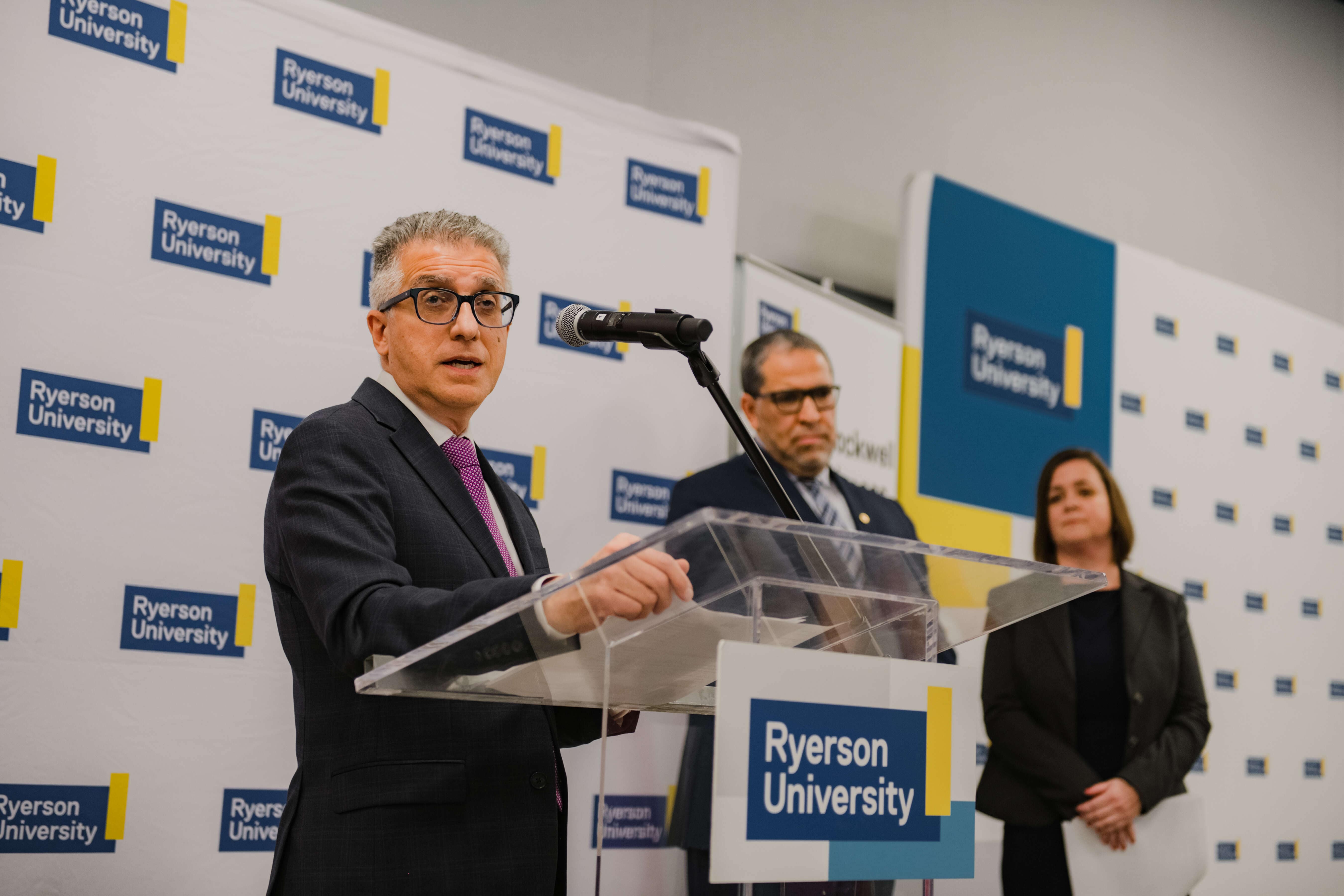 A speaker addresses an audience from a podium during a university announcement or ceremonial event. Two individuals stand behind the speaker, with university-branded signage displayed in the background.