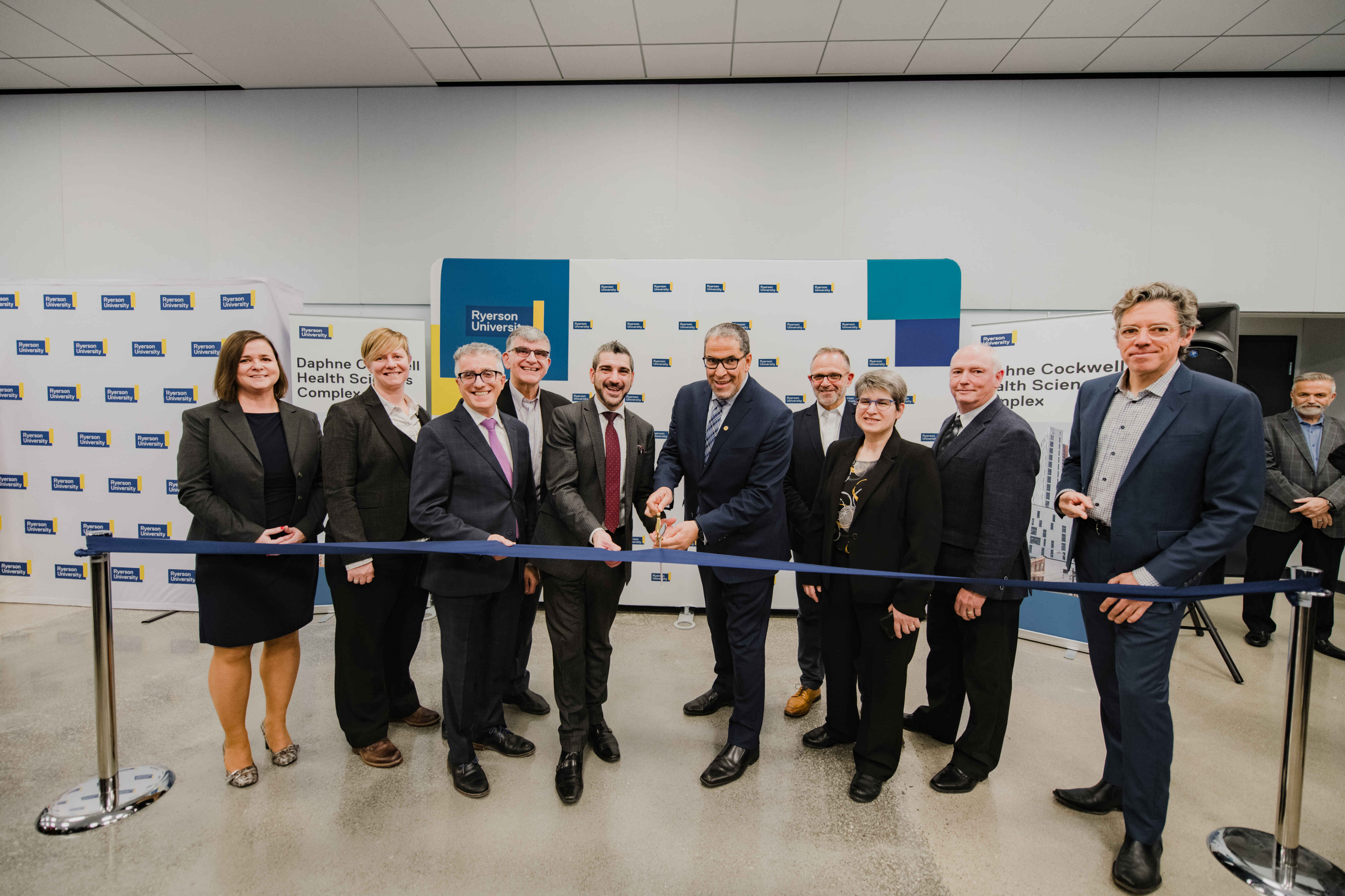 A group of university leaders and partners gather for a ribbon-cutting ceremony in front of university-branded displays. Several participants stand together smiling as the ribbon is officially cut to celebrate the opening of a new facility.