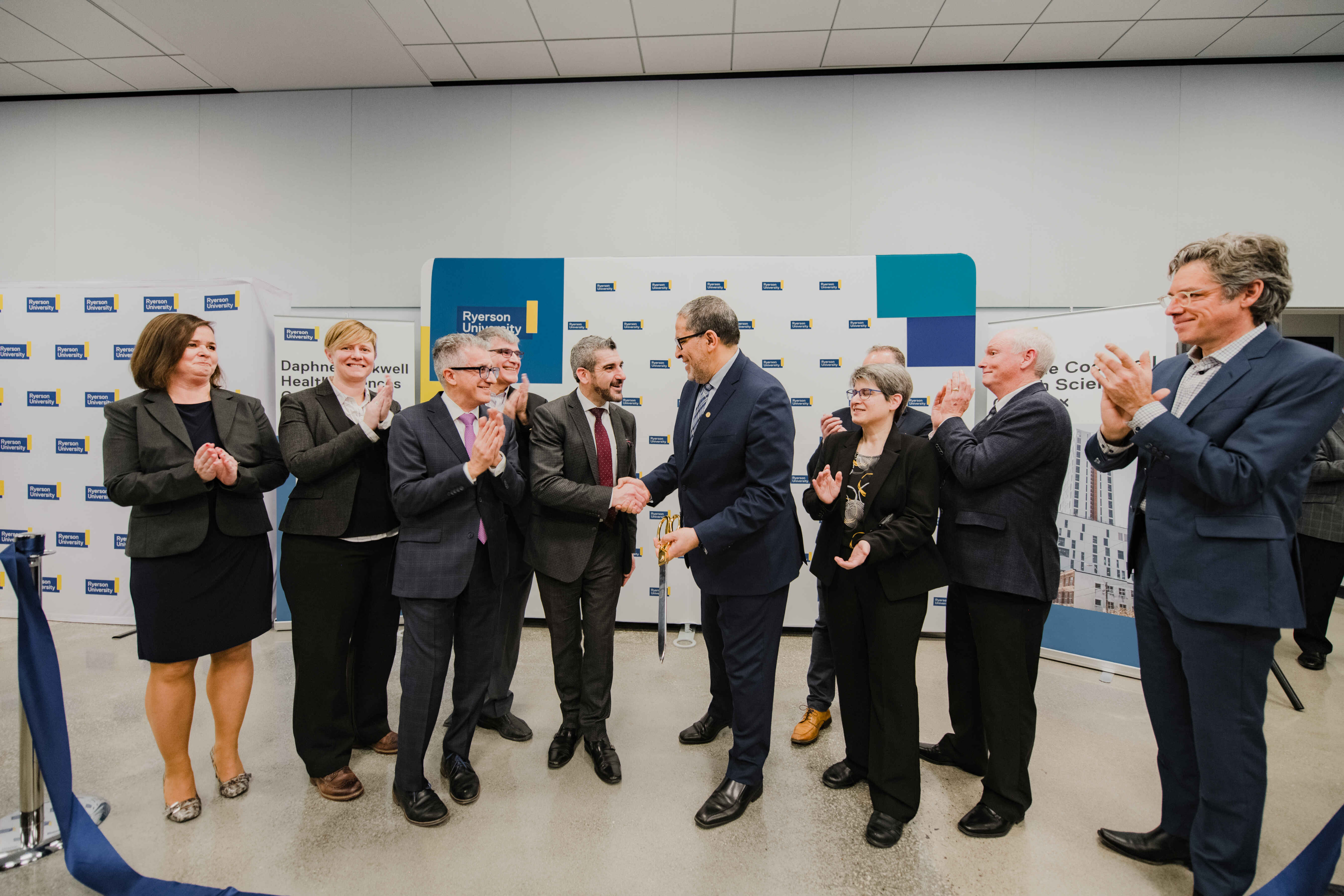 Participants applaud and shake hands following a ribbon-cutting ceremony marking the opening of a new university facility. Event attendees stand nearby celebrating the milestone.