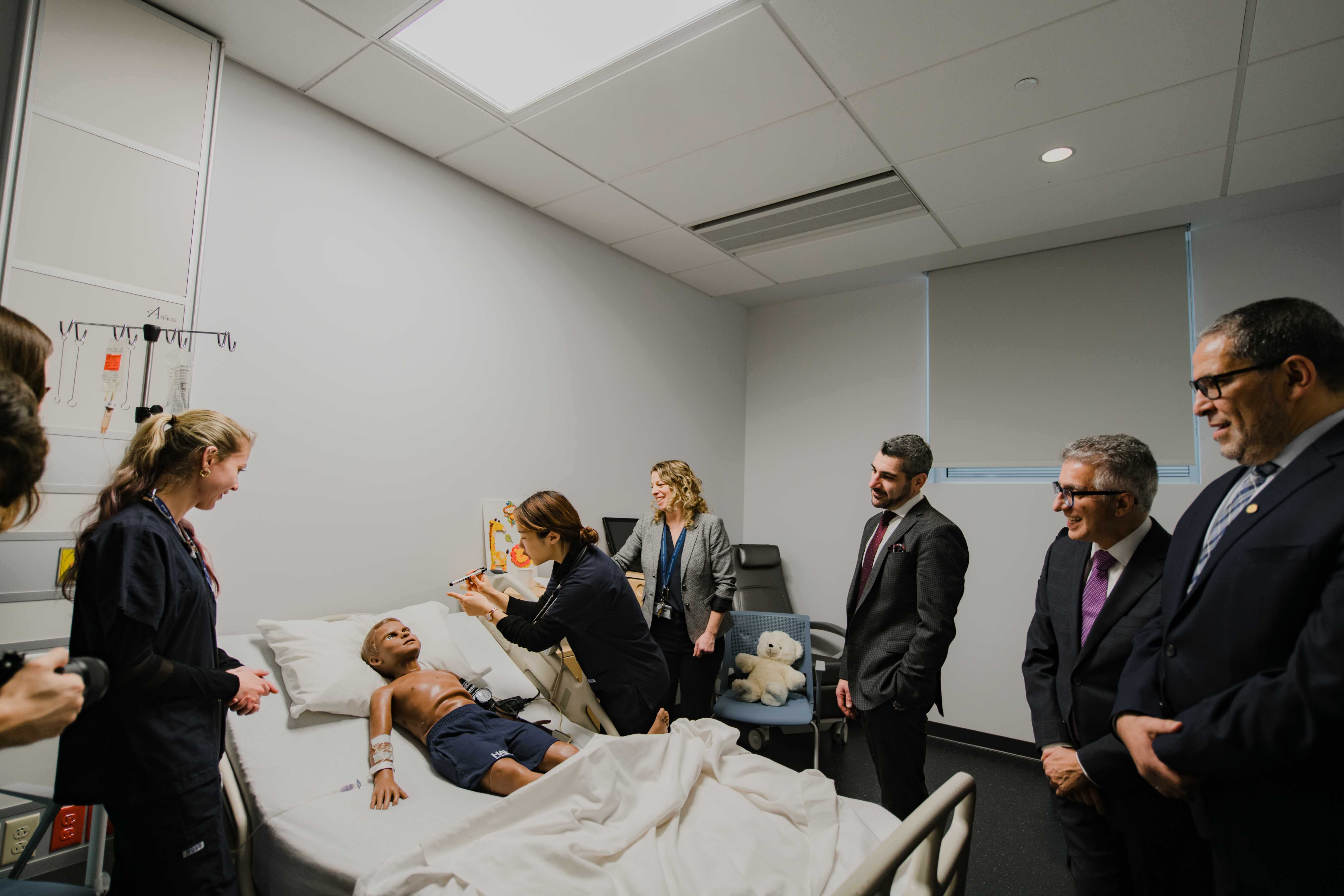 Students and visitors gather in a healthcare simulation room during a guided tour. A student demonstrates equipment beside a patient mannequin while attendees observe the learning environment.