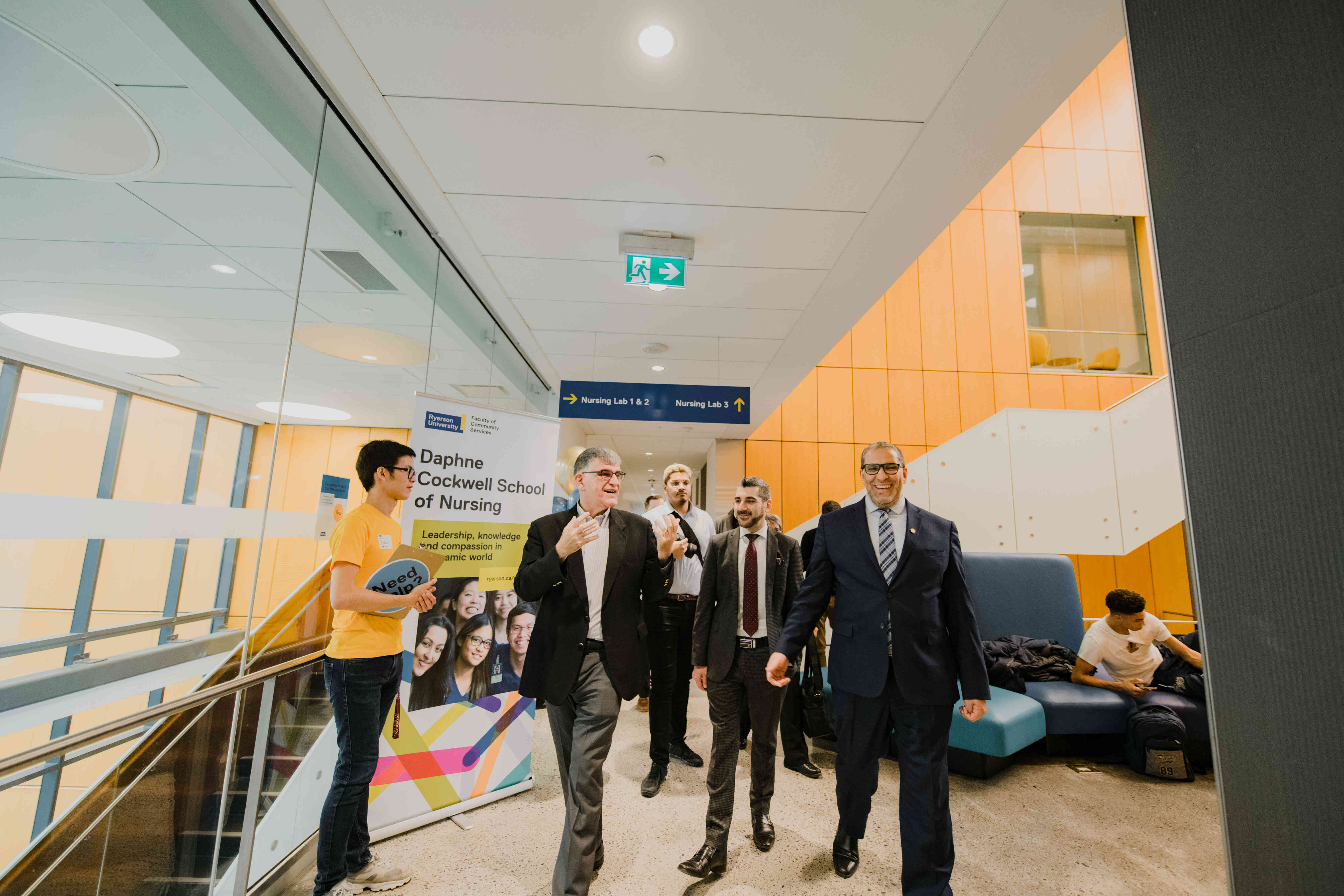 A group of university leaders and guests walk through a bright academic building hallway during a campus tour. School of Nursing signage and student information displays are visible in the background.