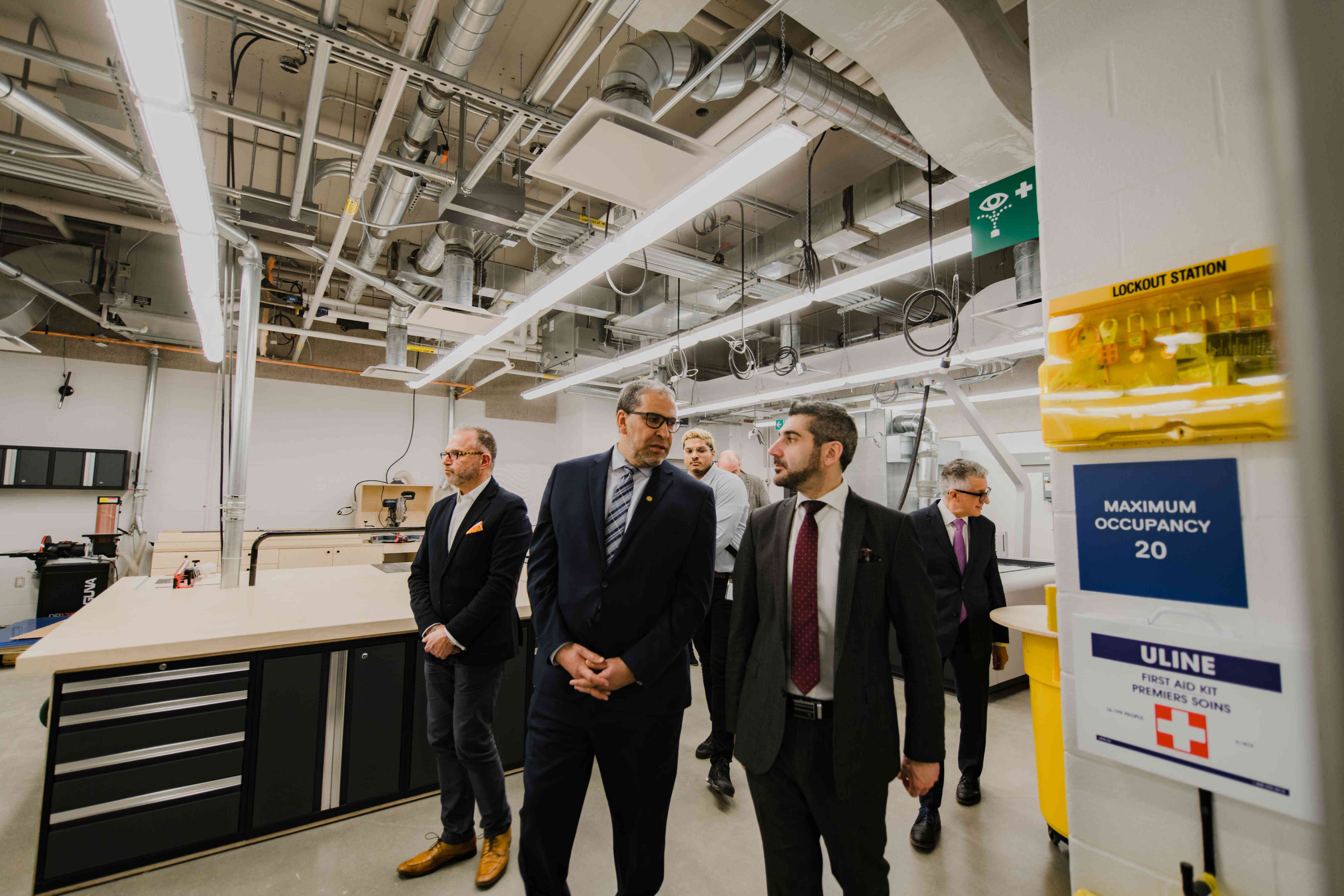 University leaders and guests tour a modern engineering and manufacturing workspace equipped with industrial tools, workstations, and safety equipment. Participants walk through the facility while discussing its features.