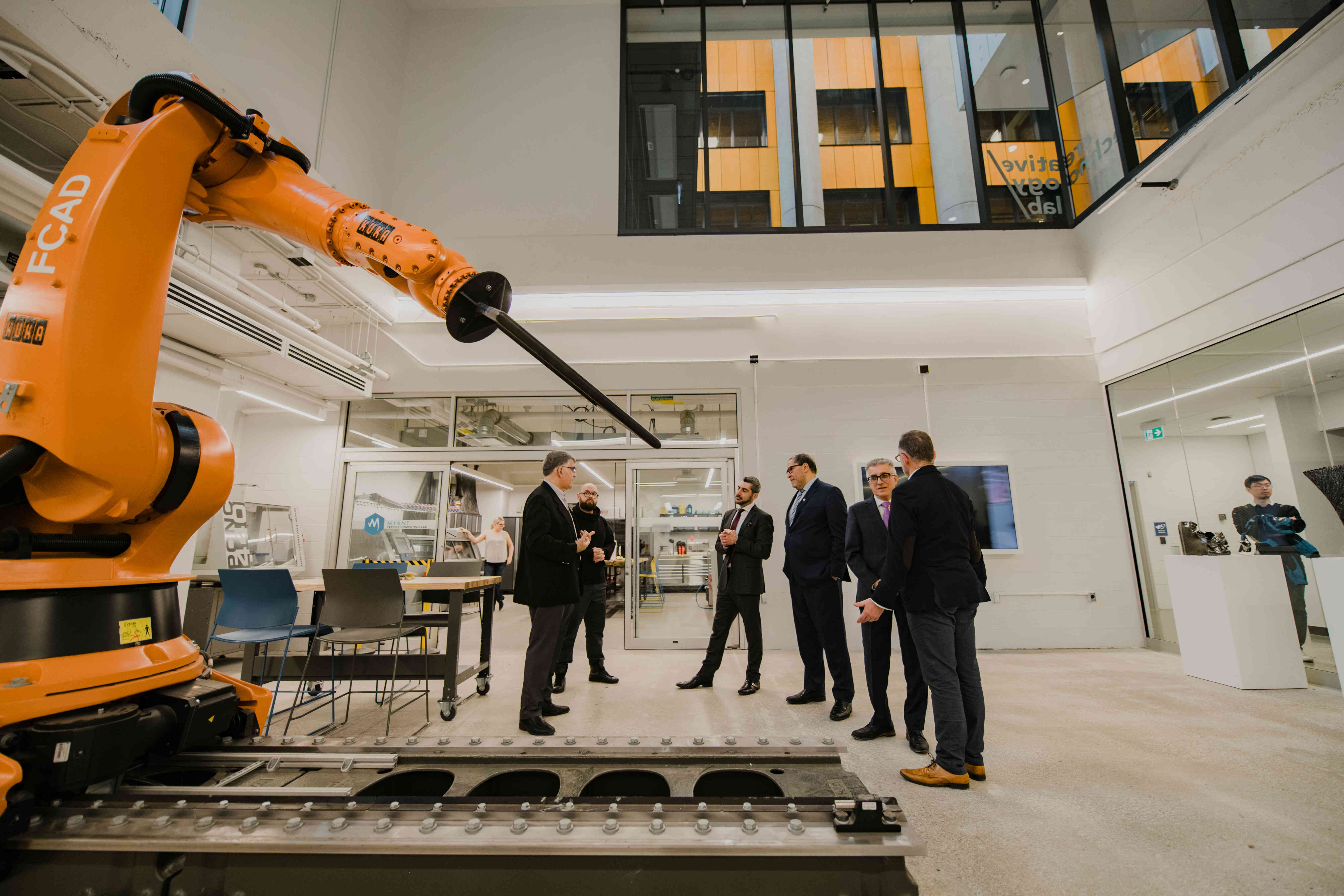 Visitors and university representatives stand in a technology and innovation lab featuring industrial robotics equipment. A large robotic arm is visible in the foreground as the group discusses the facility and its capabilities.