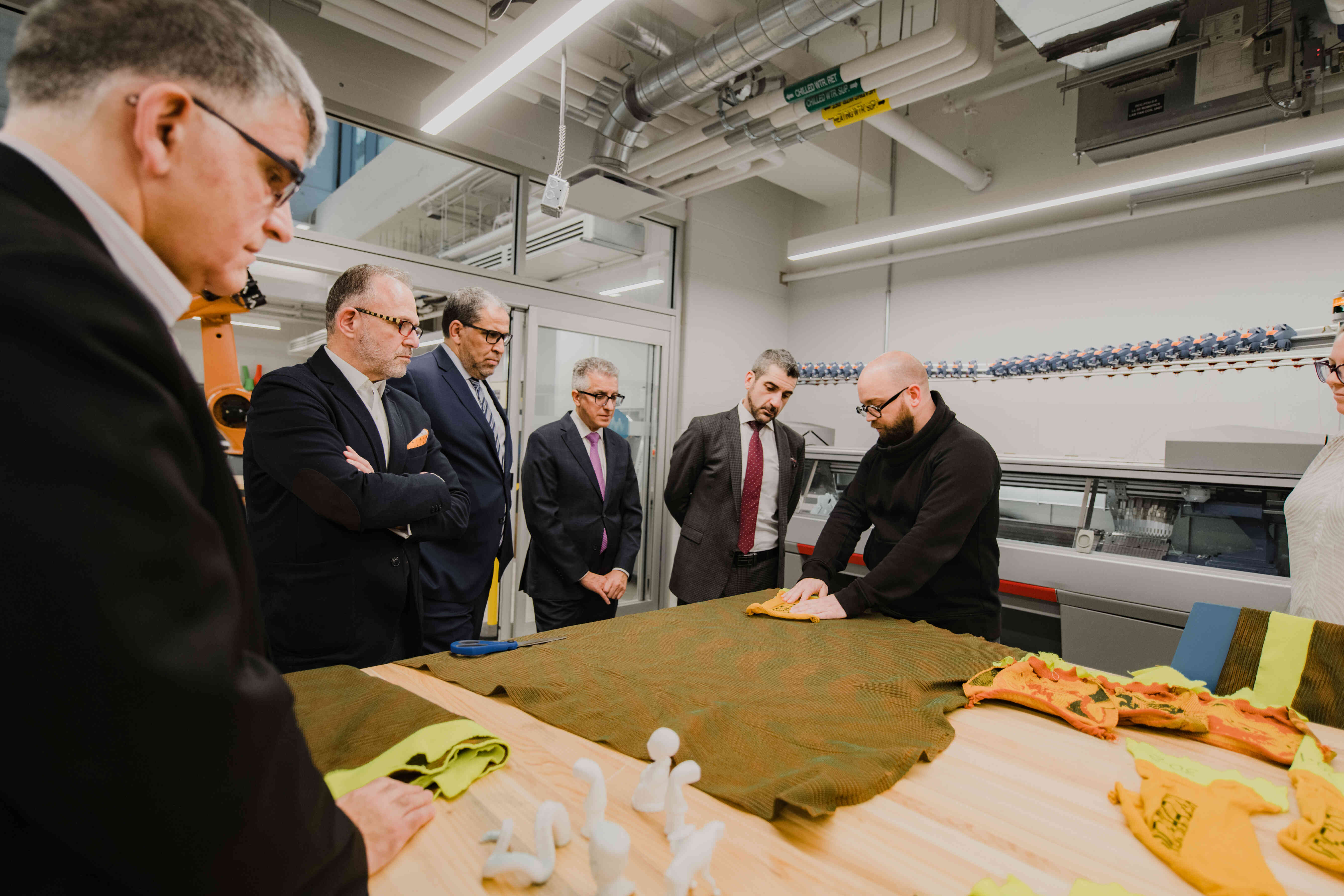 Visitors observe a demonstration in an advanced fabrication and design lab. A presenter showcases materials and equipment while guests gather around a worktable to learn about ongoing projects.