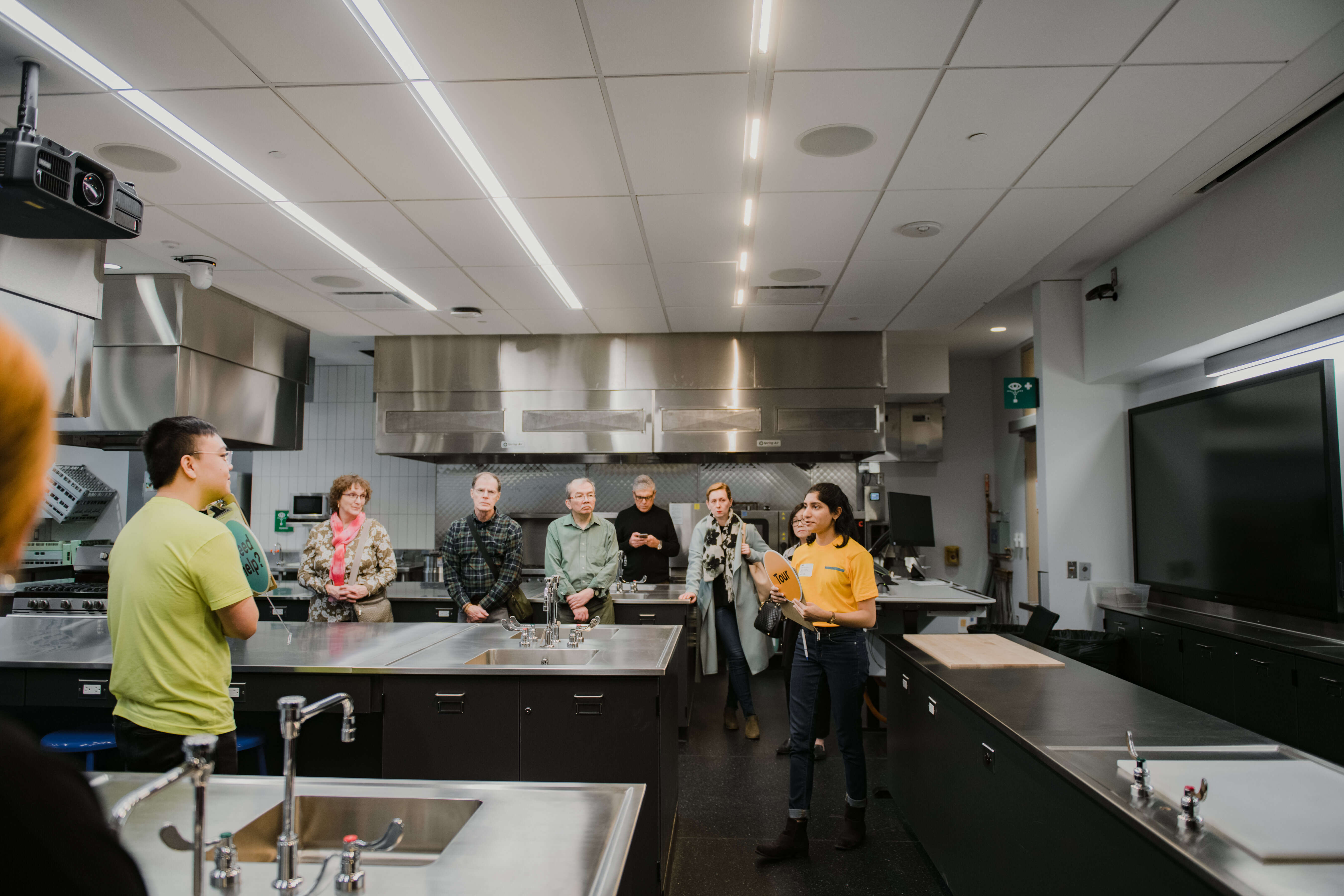 Guests participate in a guided tour of a modern teaching kitchen and laboratory space. A student guide speaks to attendees while they observe the room’s equipment and learning areas.