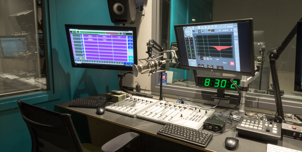 Empty Allan Slaight Radio Institute studio with a soundboard, two computer monitors, a microphone, and a wall clock reading 8:30.