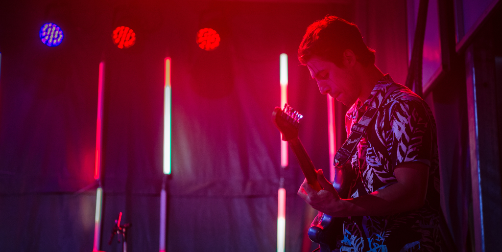 A musician in a patterned shirt plays an electric guitar on stage, bathed in red and purple lighting with vertical LED lights behind him.