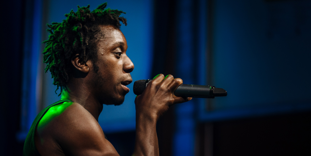 A close-up side view of a performer holding a microphone and singing under stage lights, with green light reflecting on their face.