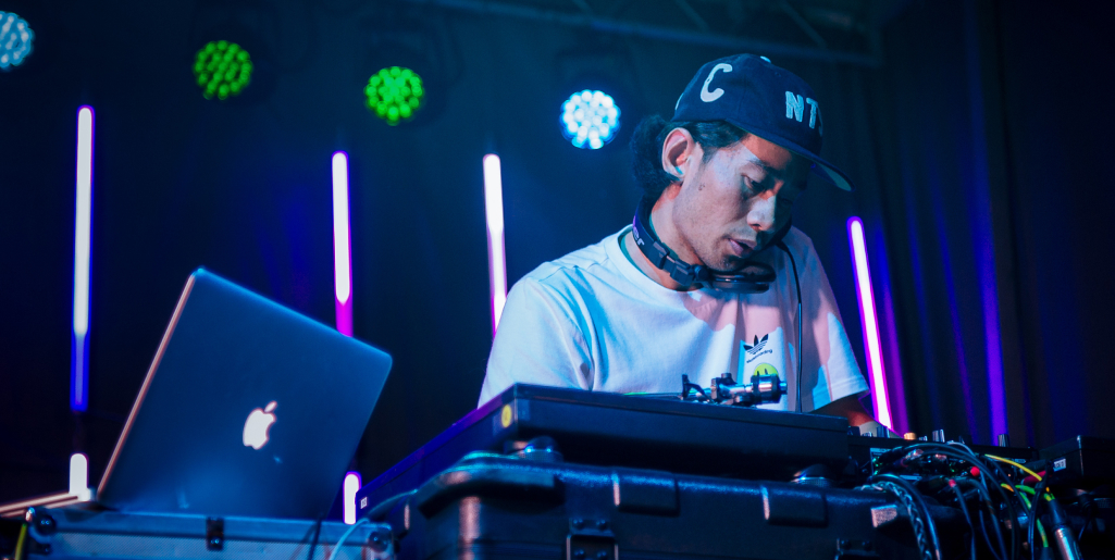 A DJ wearing a cap and headphones leans over turntables, mixing music with colorful stage lights in the background.