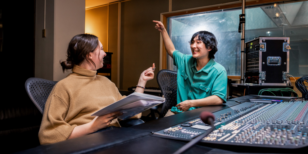Two students sit in a recording studio, smiling and pointing while discussing work at a soundboard with microphones and equipment.