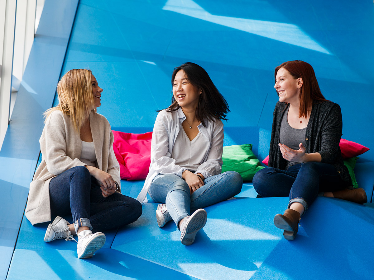 Three students laugh together as they sit in a sunlit section of the SLC. 