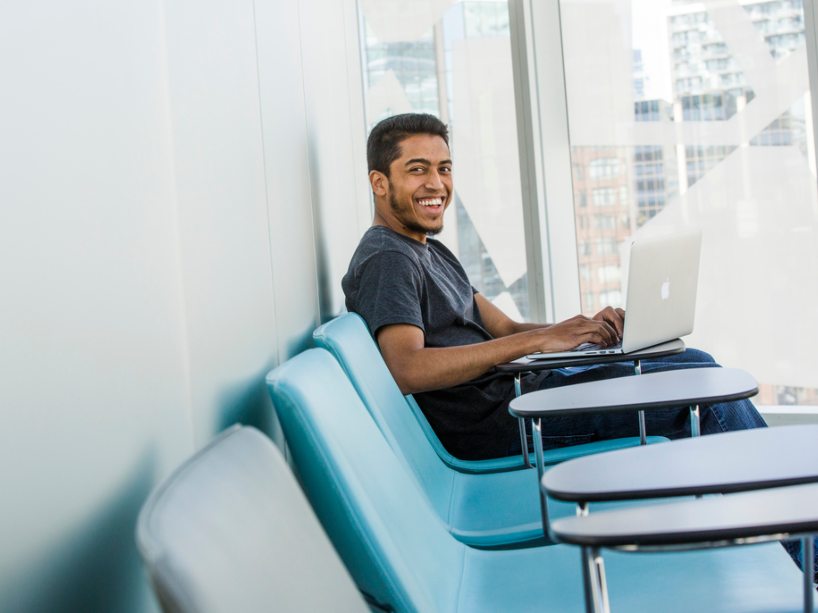 A male student sits on a blue chair and smiles at the camera.