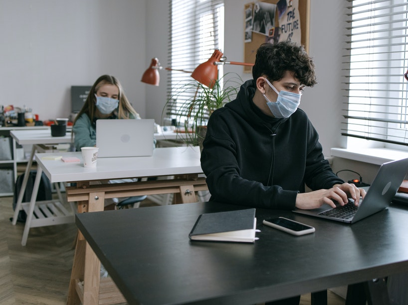 A male and female student work on laptops in a classroom while wearing masks.