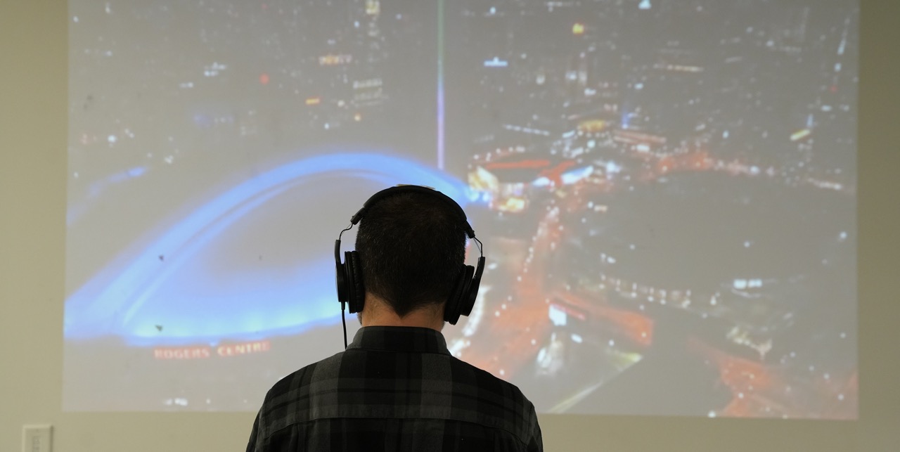 A student stands in front of a wall with a projection of the toronto skyline, they are wearing headphones
