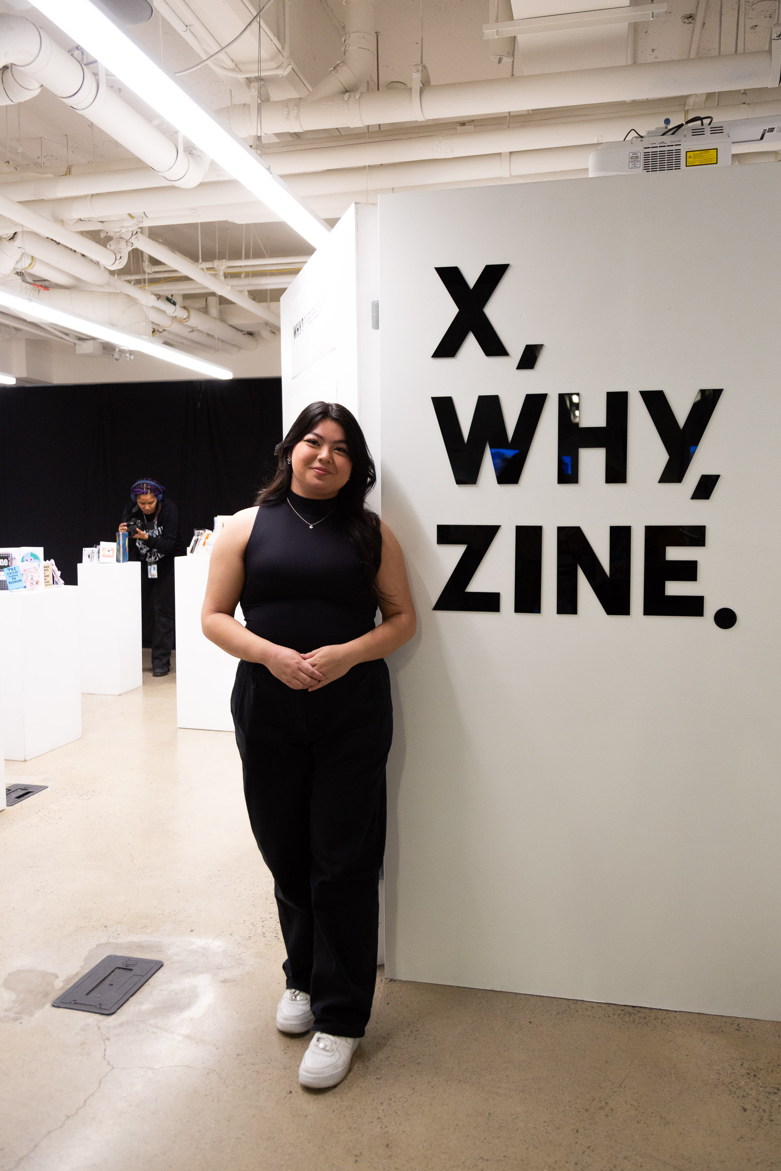 Madeline stands in front of a white wall with black text that reads X Why Zine 