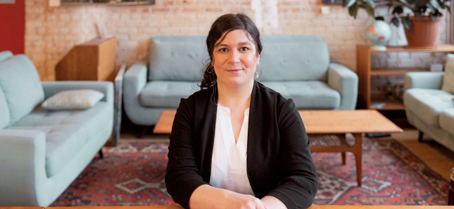 Desneige Frandsen in a black blazer and white top sits at a wooden table, smiling at the camera in a cozy lounge with blue upholstered sofas, a patterned rug, and brick walls behind them.