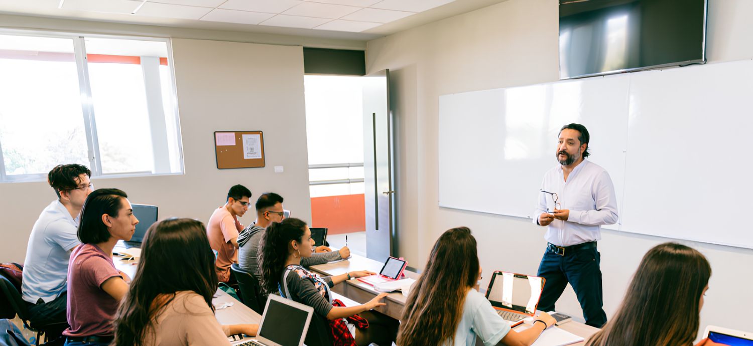 A trainer stands at the front of a bright classroom, speaking to a group of people seated at desks with laptops, while one person writes or gestures during a workshop.