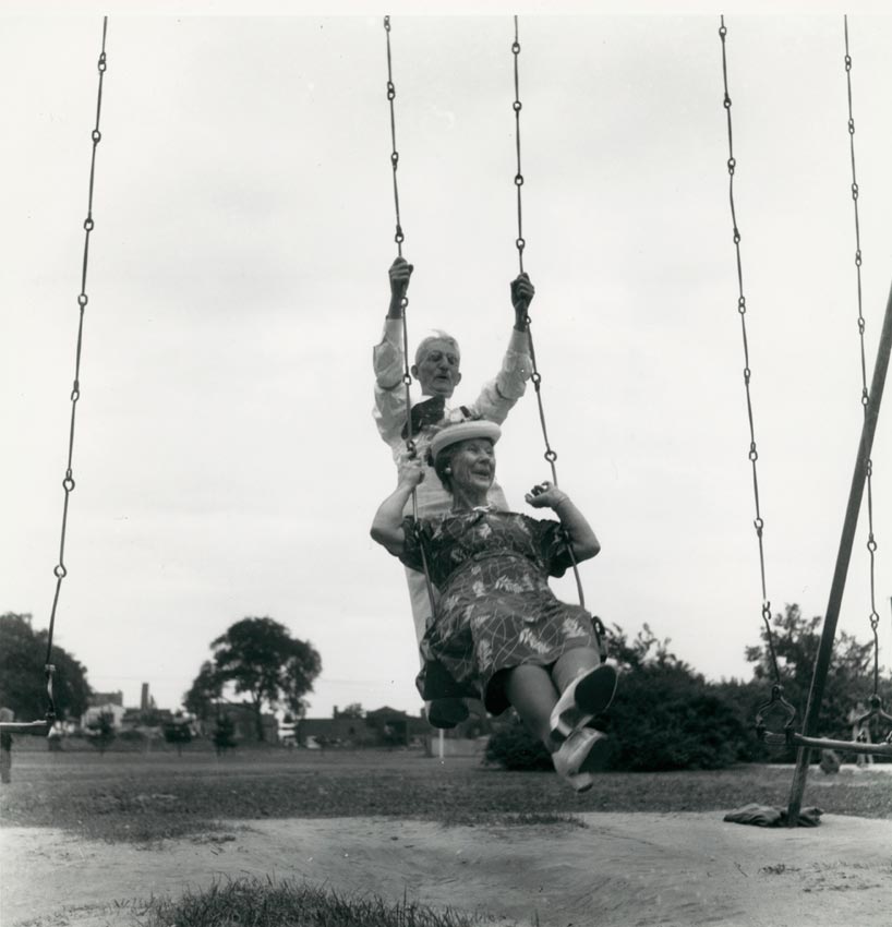 Black and white photo of an elderly man pushing an elderly woman on a swing