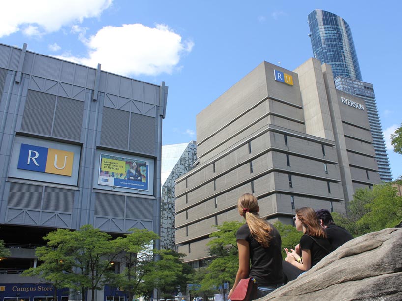 Three females sitting on rocks near Lake Devo facing Victoria St. garage