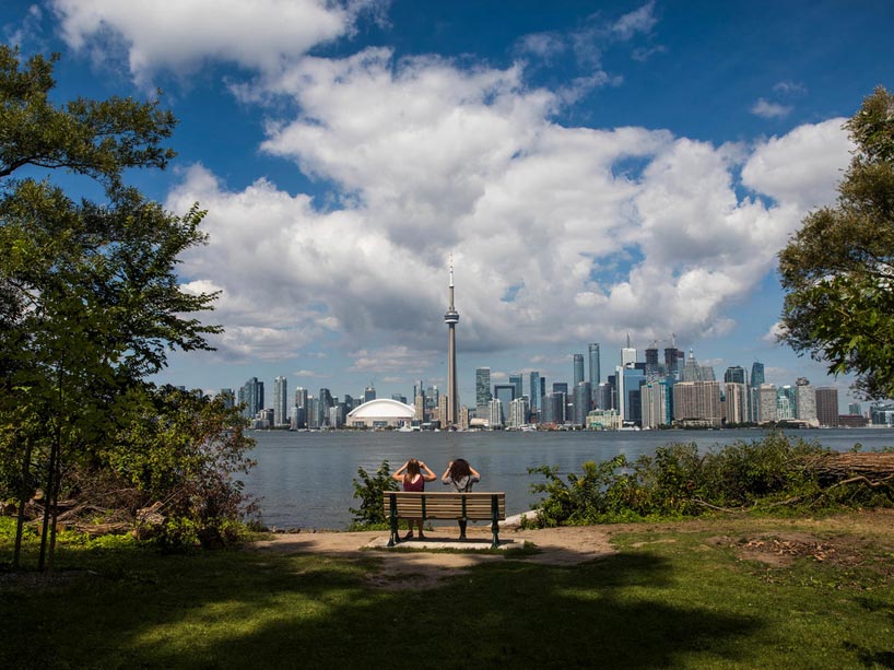 Two people sitting on a bench using binoculars to look across water at the city of Toronto skyline