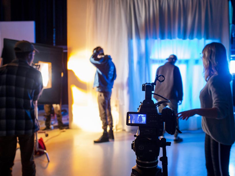 One student, left, holds a light on two actors; a fourth student, right, stands by a camera