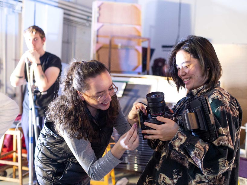 Two students hold a camera, adjusting the bottom of it, while a third student looks on in the background