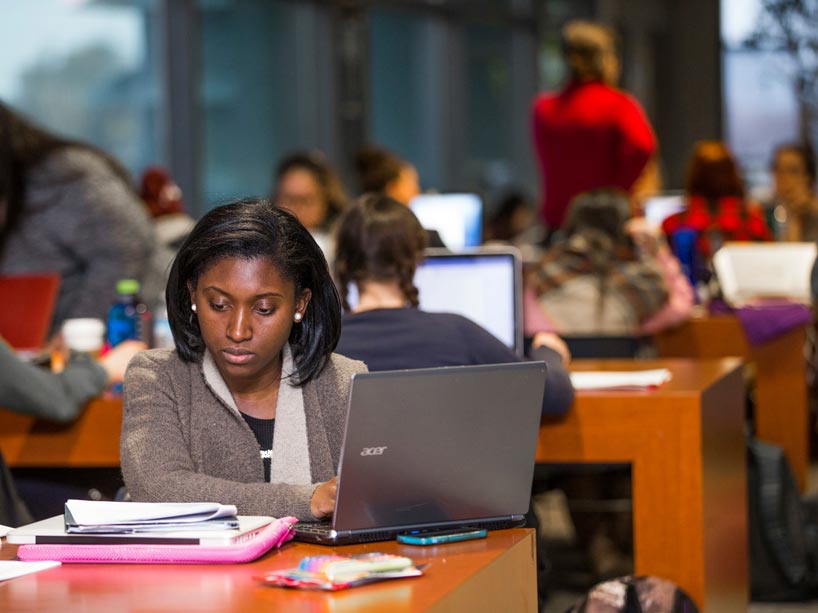 A young woman typing on her laptop with students in the background studying