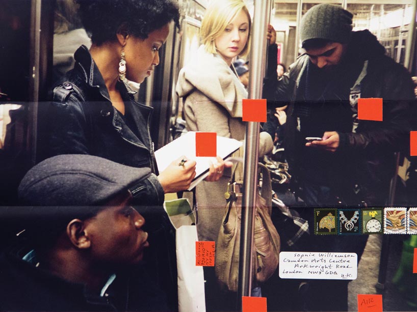 Two women and two men ride a crowded subway car, one of the women is writing in a notebook, while one of the men looks at his phone screen.